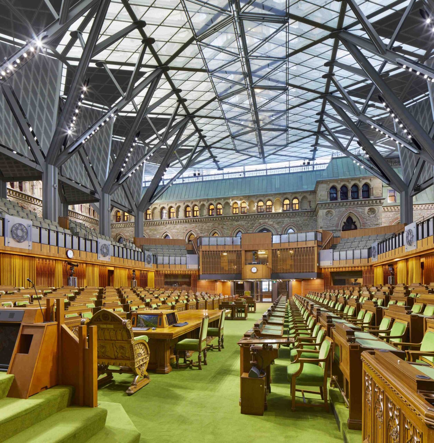 The interior of the interim House of Commons Chamber with the sun shining through the vaulted glass ceiling. The Chamber has green carpet and green upholstered chairs.