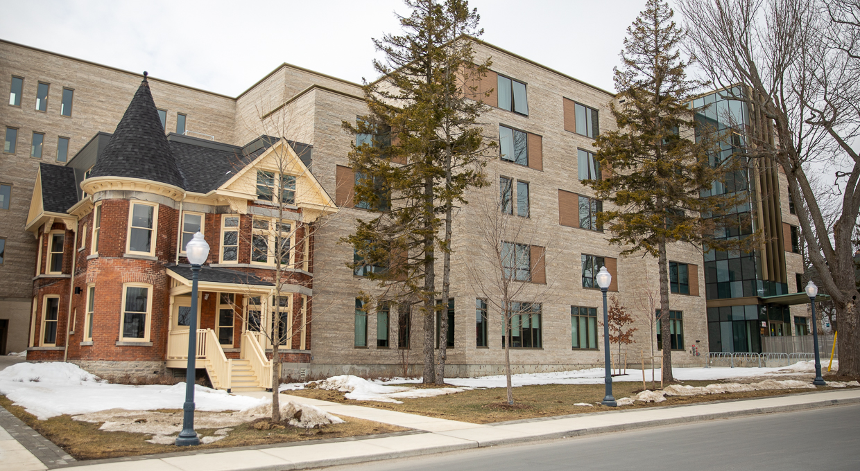 The image shows the Queen's University Endaayaan – Tkanónsote residence, a modern building, light beige stone-clad, with large windows, adjacent to a smaller, older brick Victorian-style house. The older house features a steeply pitched roof, a prominent turret, and decorative detailing.