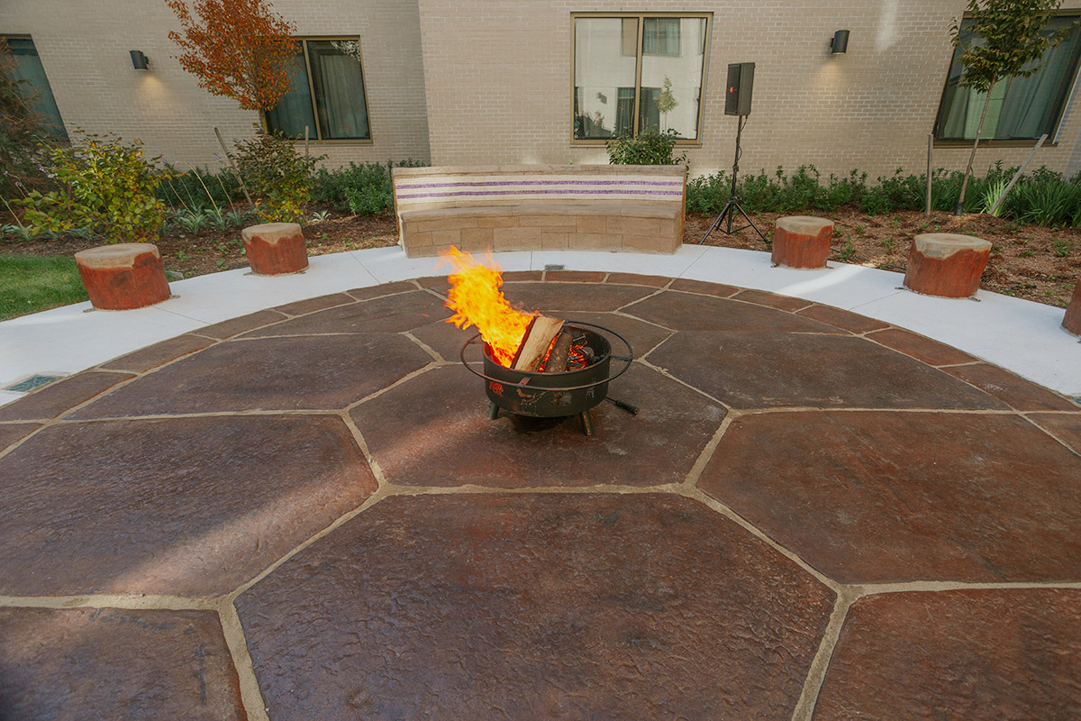 A circular courtyard area outside Queen’s University’s Endaayaan – Tkanónsote residence, paved with large, dark reddish-brown, irregularly shaped stones. A metal fire pit with burning wood sits in the center of the patio. Small, log-shaped, light-colored wooden stools are placed around the perimeter. A light beige/tan colored building with windows is visible in the background. Various plants and shrubs are visible in the landscaping around the patio. A wooden bench is also visible.