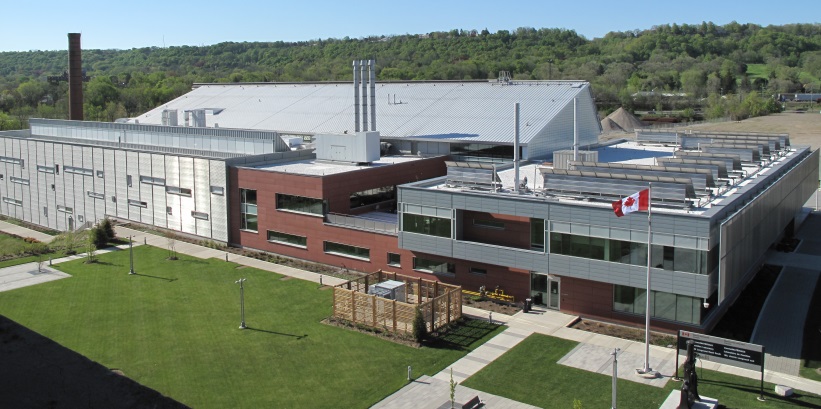 The image shows an aerial view of a modern, multi-building complex situated on a landscaped lawn. The main structure features a large, flat-roofed building with metal siding, which functions as a laboratory. A smaller, brick building is attached. A Canadian flag is visible.