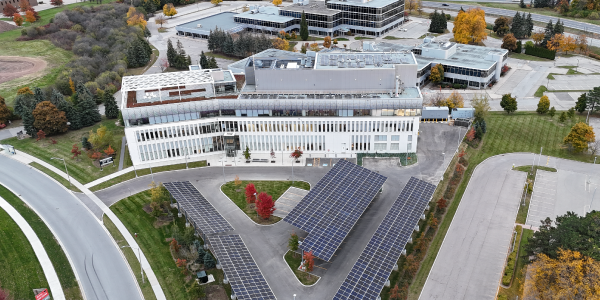 The image shows an aerial view of a modern, light-colored research facility, situated within a landscaped campus. The building features a flat roof and extensive window lines. In front of the building, there's a sizeable solar panel array forming a V-shape. The surrounding area is well-maintained, with paved roads, grassy areas, and autumnal trees showcasing vibrant fall colors.