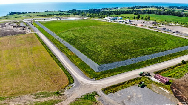 Aerial view of the Port Granby Long-Term Waste Management Facility, a large, grassy rectangular field bordered by roads, with buildings, farmland, and a body of water in the background.
