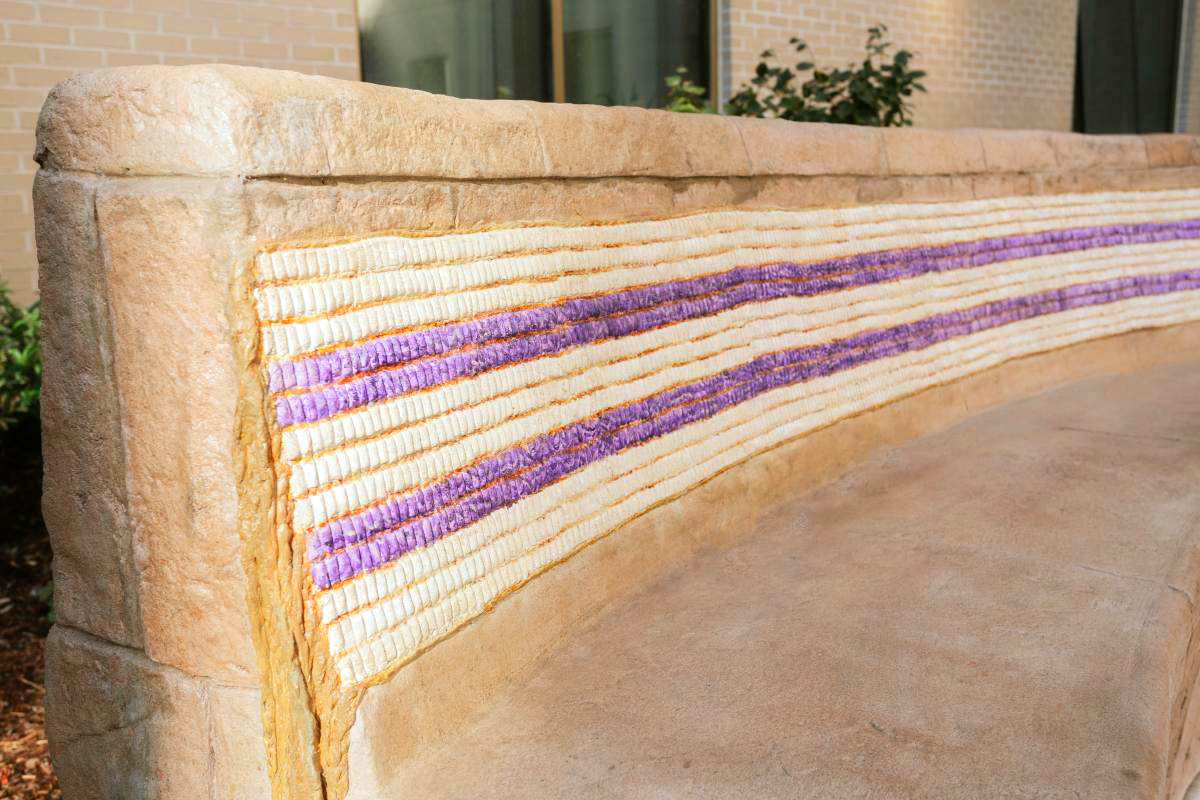A curved, light beige-tan stone bench that represents the Two Row Wampum belt pattern. The back of the bench is decorated with a mosaic-like design. Small, off-white and light purple, almost lavender colored, beads or tiles form horizontal bands. These bands of color appear to be laid in a pattern that resembles woven stripes across the back of the bench. The pattern is consistent across the curvature of the bench back. The stonework appears to be a natural stone, with a slightly rough or textured surface. The bench’s edges are beveled, and the back appears slightly thicker than the seat area.