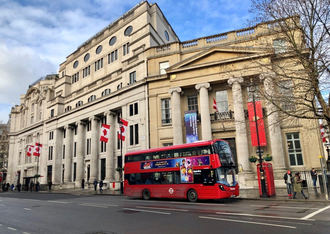 A red double-decker bus navigates a city street, flanked by the Canada House building and pedestrians.