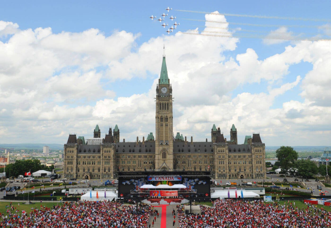 Centre Block during Canada 150 celebrations. A grand structure with a distinctive clock tower rising above its roofline. A large crowd is gathered in front of the building, which has a large stage setup out front.