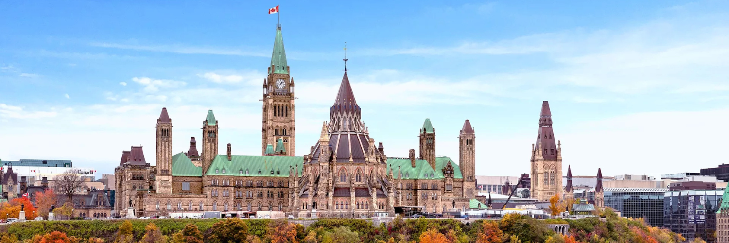 A view of Parliament Hill from a distance, with the tower of Centre Block visible. The photo was taken in the fall so trees with changing colours are visible in the foreground.