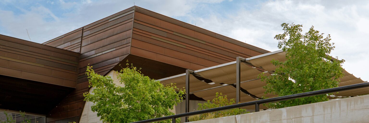 A modern building with metal siding. A pergola with retractable shades and trees are in the foreground.