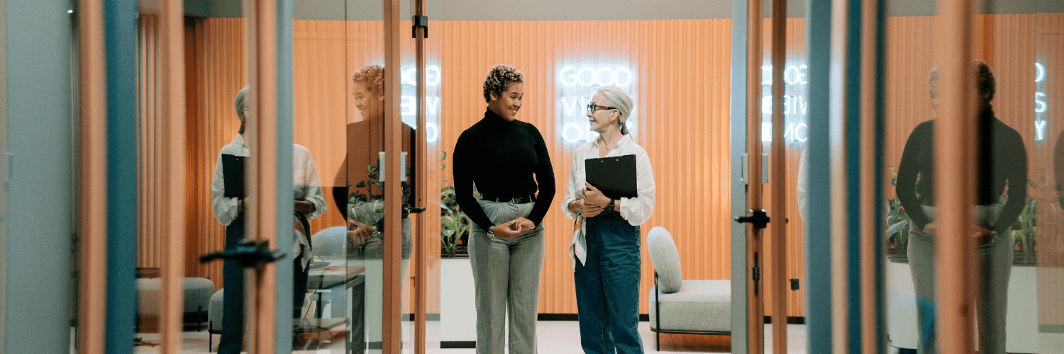 Two women standing at the end of an office hallway conversing. Doorways are visible in the hallway in front of the women.