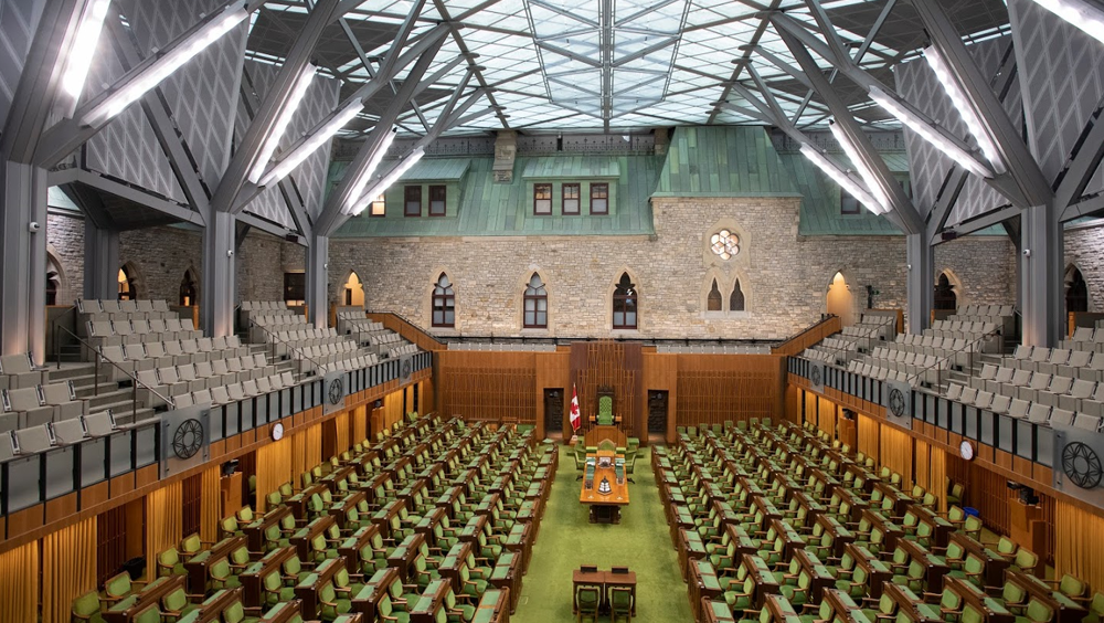 The interior of a glass-roof structure is pictured with the brick wall of a heritage building incorporated as the back wall. Seating is pictured throughout the space with green carpeting underneath.
